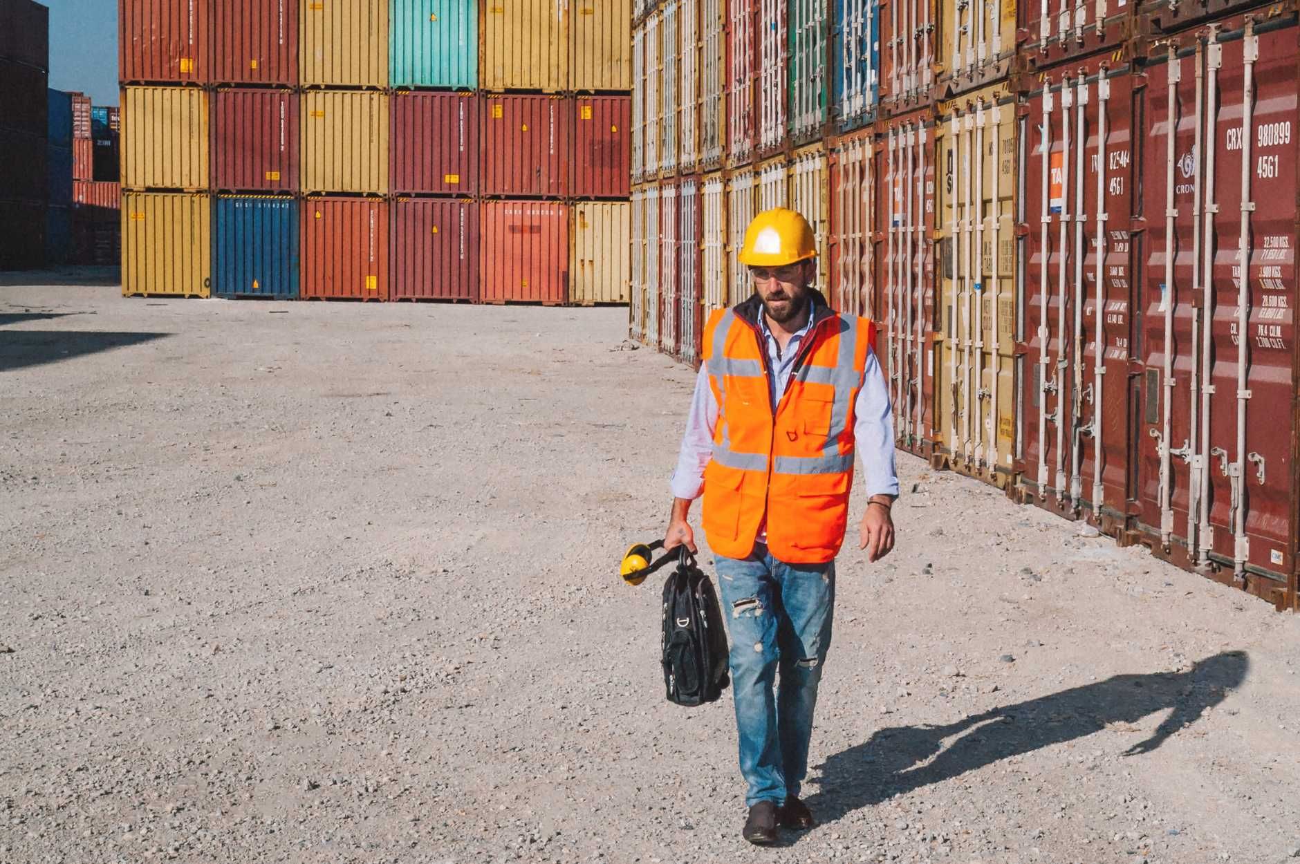 Man wearing safety vest and hard hat walking through depot with stacked containers